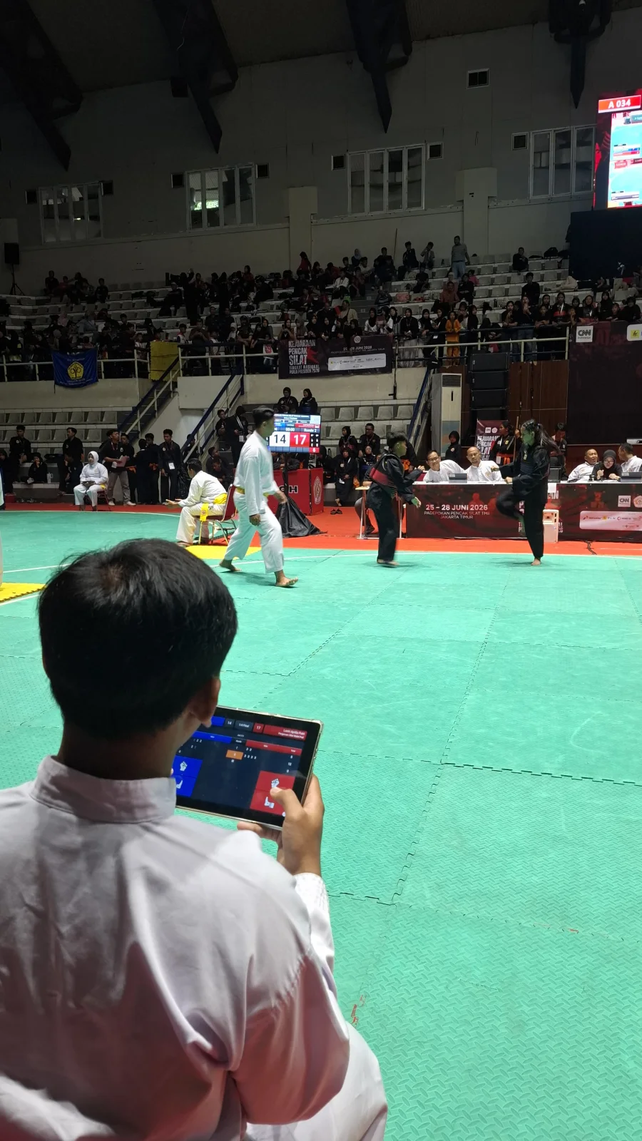 Suasana pertandingan Kejuaraan Pencak Silat CNN Indonesia 2 Piala Presiden di Padepokan Pencak Silat TMII, Jakarta Timur, dengan pesilat bertanding di atas gelanggang dan papan skor digital terlihat di arena.